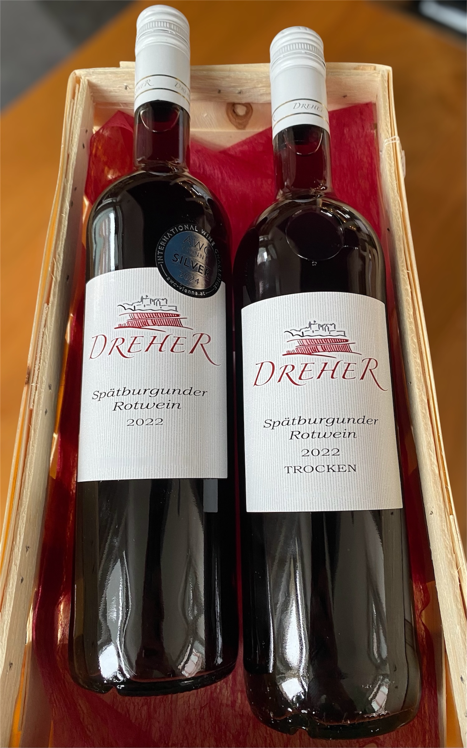 Zwei Weinflaschen Dreher Spätburgunder Rotwein in einer Holzbox auf rotem Stoff - links die feinherbe, rechts die trockene Variante. Ideal für junge Weinliebhaber:innen, leicht und fruchtig.